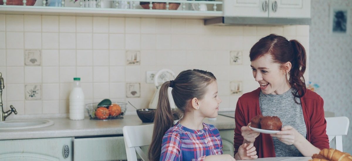 Young happy mother bringing tasty muffins to her cute daughter sitting at the table in kitchen waiting for breakfast at home