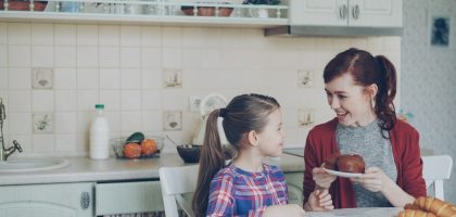 Young happy mother bringing tasty muffins to her cute daughter sitting at the table in kitchen waiting for breakfast at home
