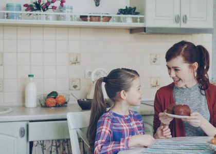 Young happy mother bringing tasty muffins to her cute daughter sitting at the table in kitchen waiting for breakfast at home